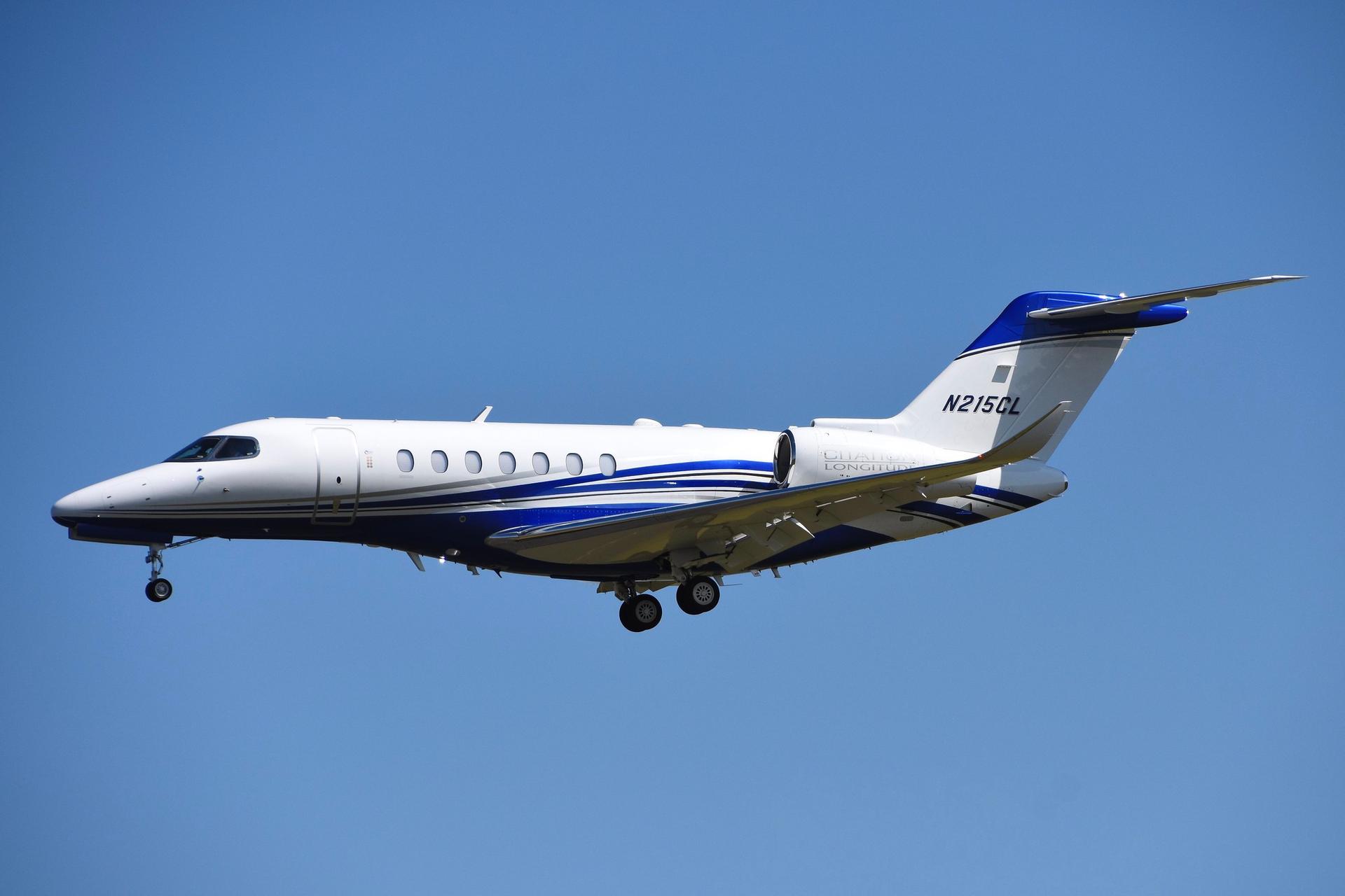 A white Cessna Citation Longitude with blue accents and a deployed landing gear in front of a blue sky.