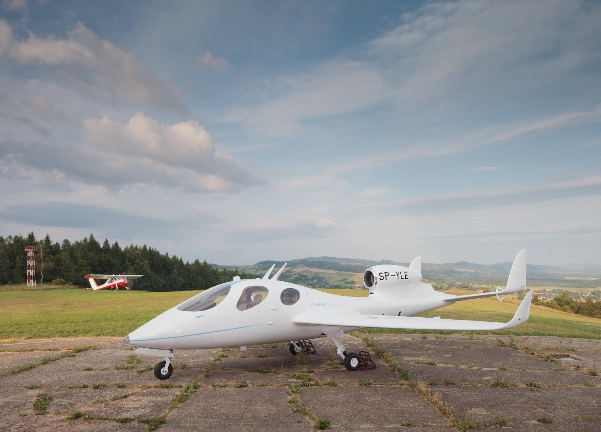 A white Flaris LAR 01 parked on the edge of a cracked concrete pad, with evergreen trees and green hills in the background.