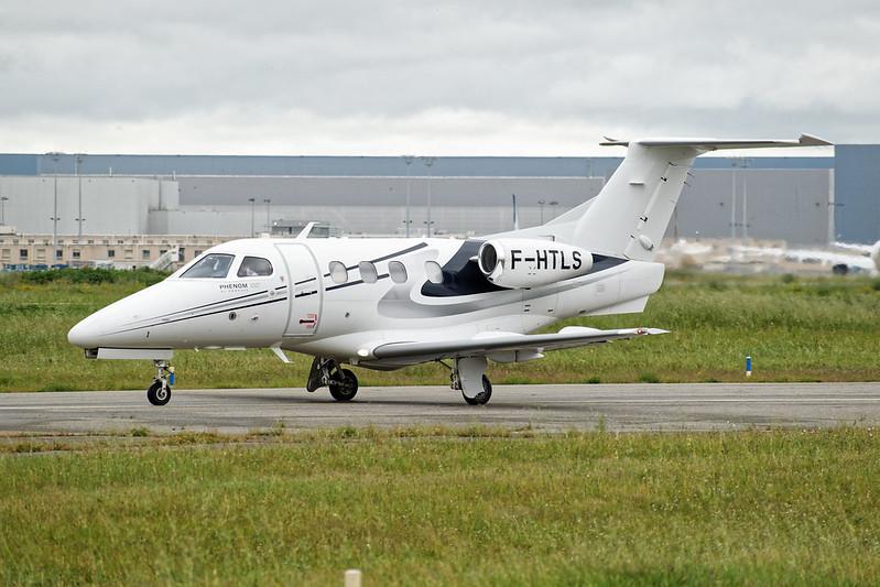 A white Embraer Phenom 100 on a taxiway, with green grass and a cloudy sky.