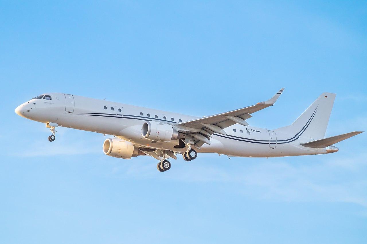 A white Embraer Lineage 1000 on approach in front of a light blue sky.