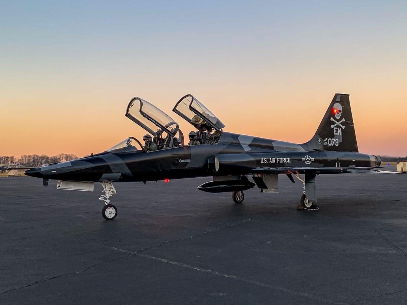 A parked T-38C Talon in a black camo with an orange sunset sky in the backdrop.