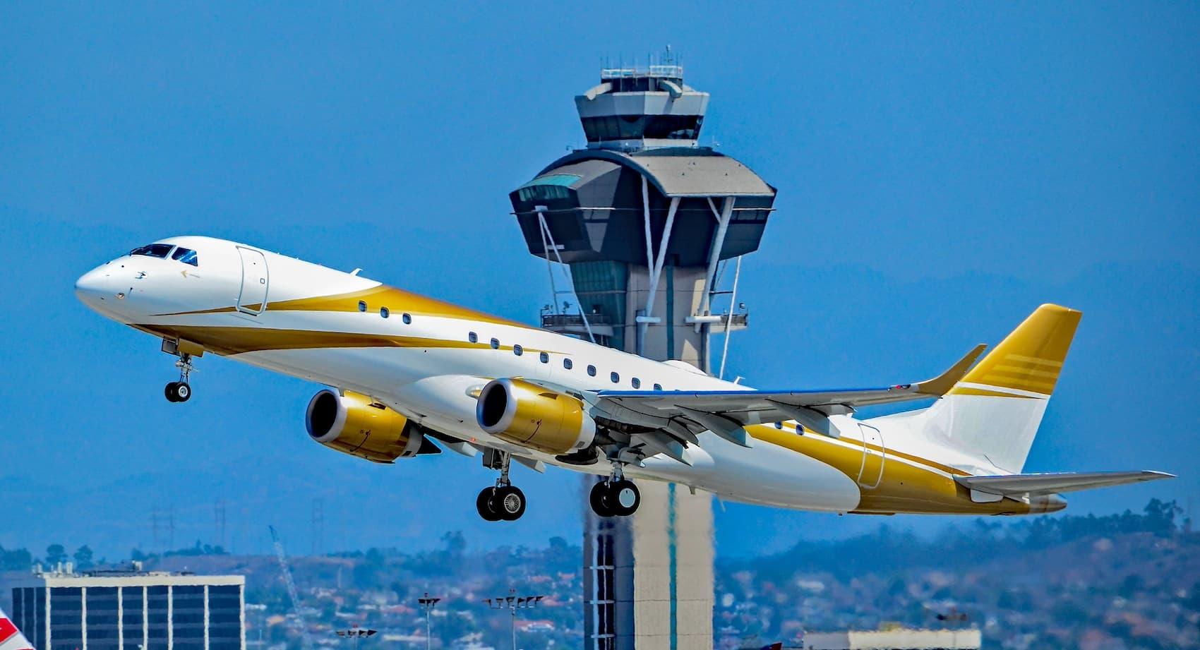 A white Embraer Lineage 1000E with gold accents taking off with a control tower in the background.